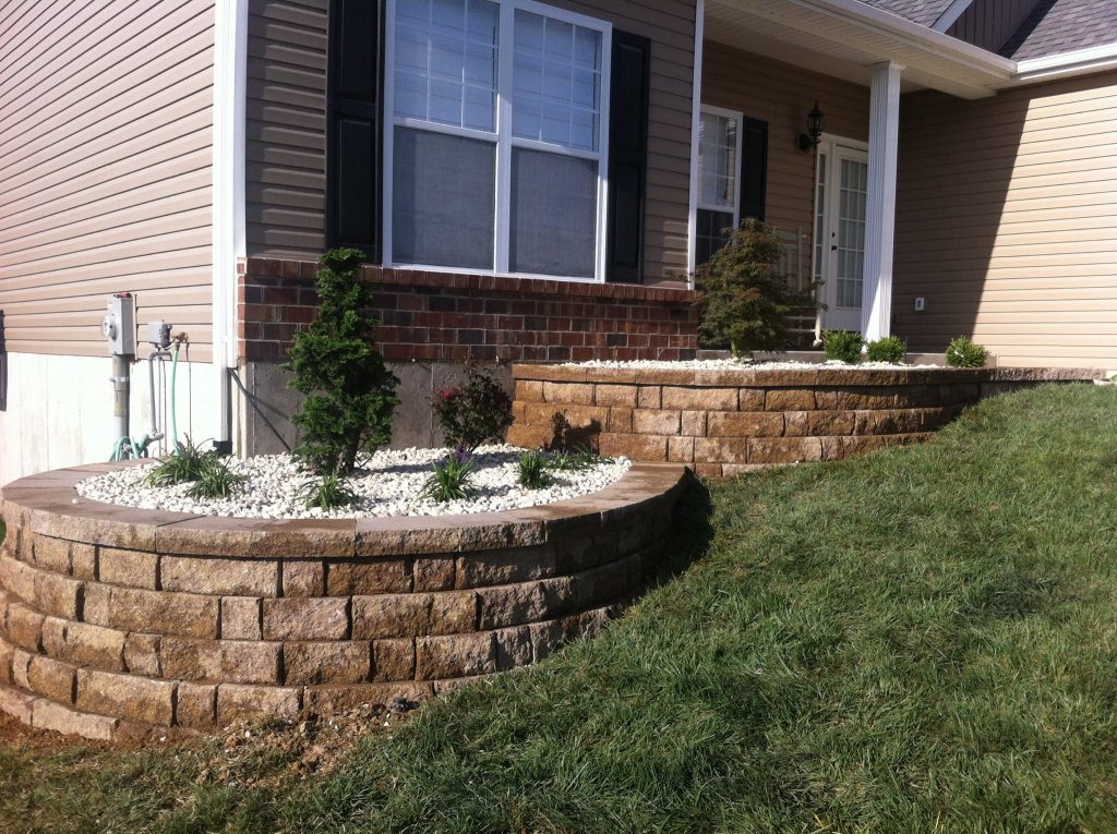 Two curved, tiered modular block retaining walls creating raised garden beds filled with white rock and small evergreens in front of a tan-siding house.
