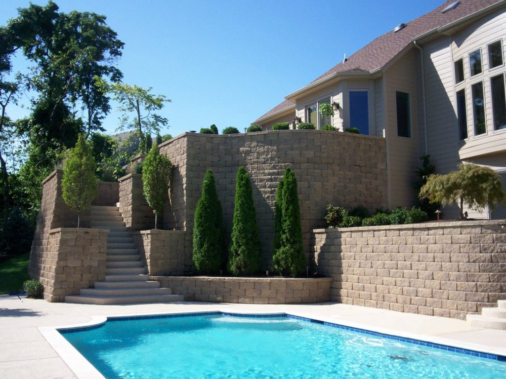 Large, tiered tan modular block retaining walls and matching stone steps ascending a slope next to a house and a bright blue swimming pool.