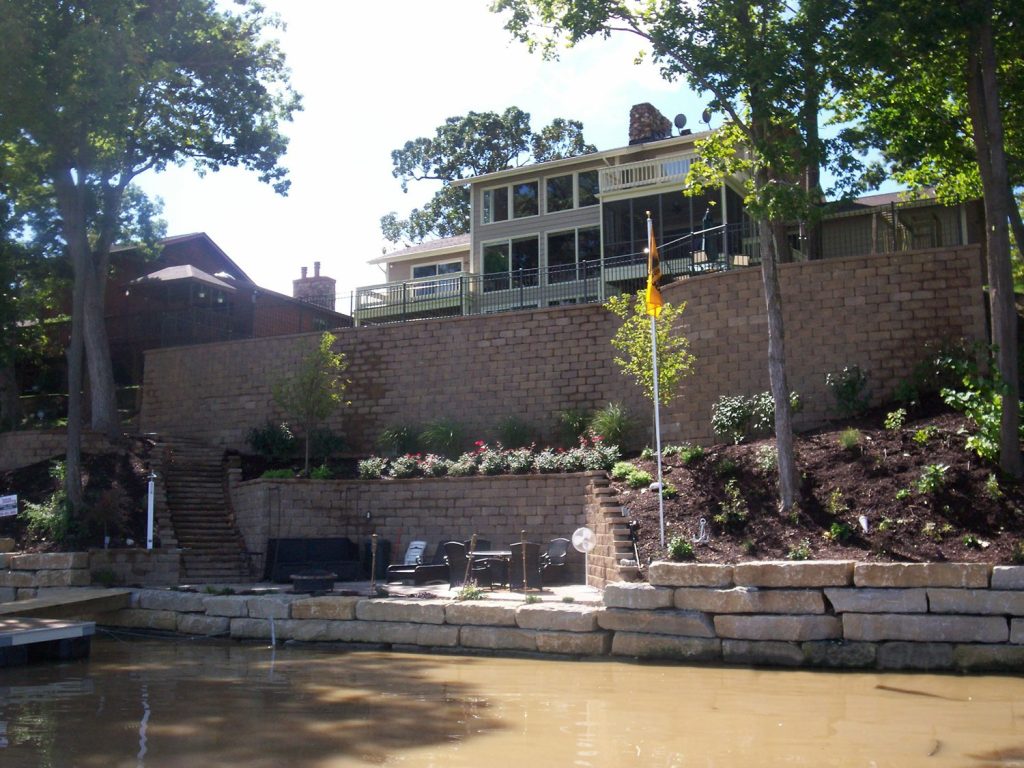 Retaining walls and steps rise from a natural stone seawall bordering a lake, up to a large house with a walkout deck.