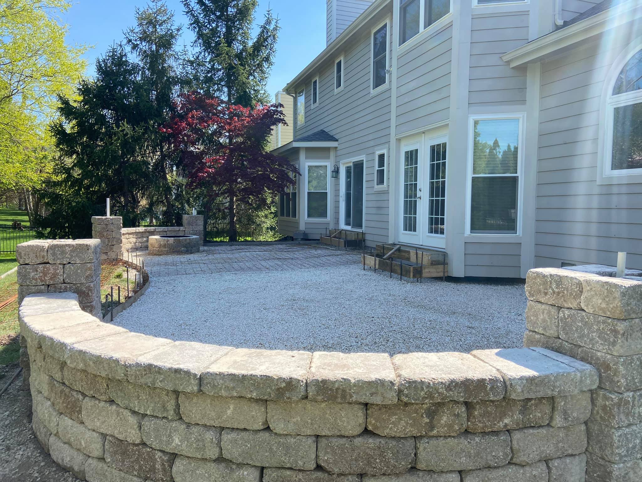 Backyard patio construction, featuring a curved retaining wall of tan modular blocks enclosing a gravel base, with a paver landing area and fire pit visible behind it.